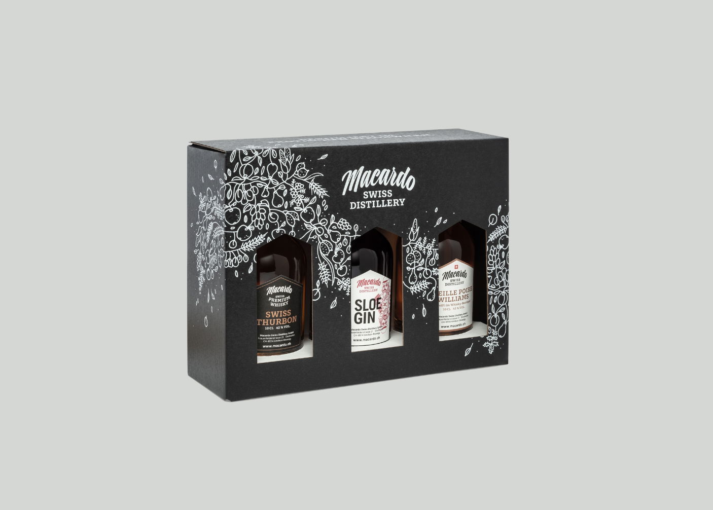 Schwarze Geschenkbox mit drei Macardo Swiss Distillery Spirituosenflaschen. Die Box ziert weiße Pflanzenornamentik und den Markennamen. Die Flaschen sind mit "Swiss Thurbon", "Sloe Gin" und "Eille Poire Williams" beschriftet.